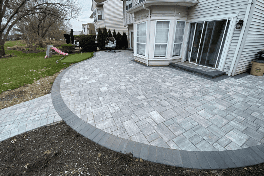 Backyard patio with gray stone paving leads to a house with sliding glass doors. A small playground slide and artificial turf are visible on the left side.