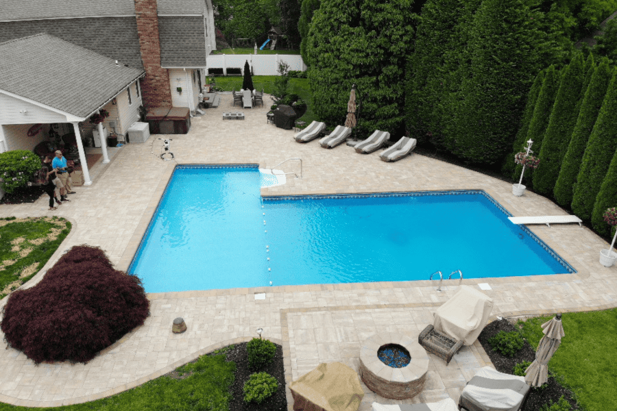 Aerial view of a rectangular swimming pool in a backyard surrounded by a patio. Lounge chairs, umbrellas, and a fire pit are nearby. House and trees in the background.