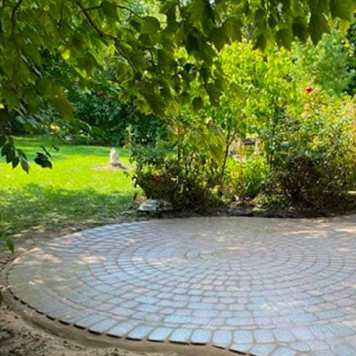 Circular stone patio in a lush garden shaded by green leaves, with a blurred view of a statue in the background.
