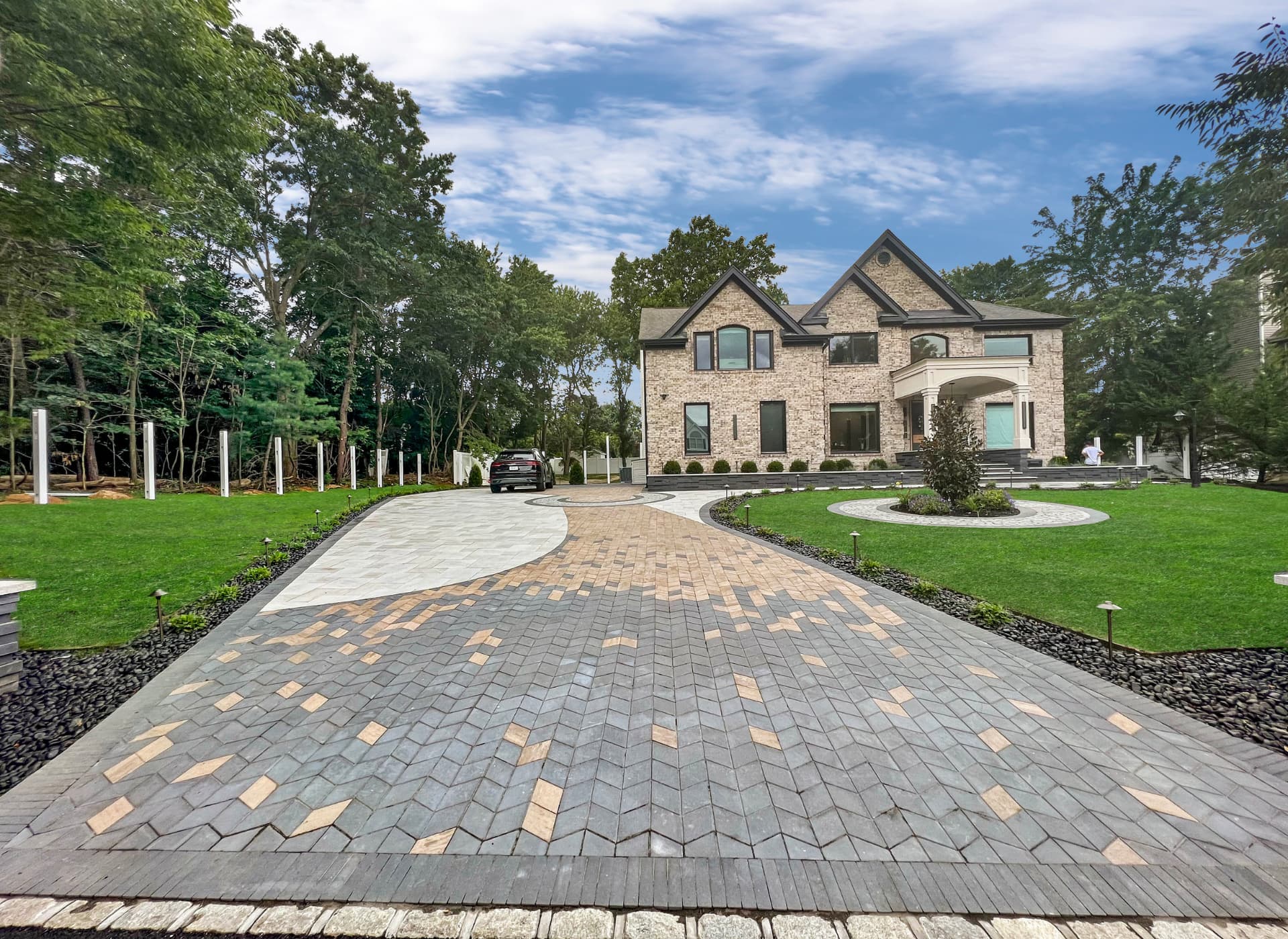 A large stone house with a spacious, patterned driveway, surrounding greenery, and trees. A black car is parked near the left side of the property. The sky is partly cloudy.