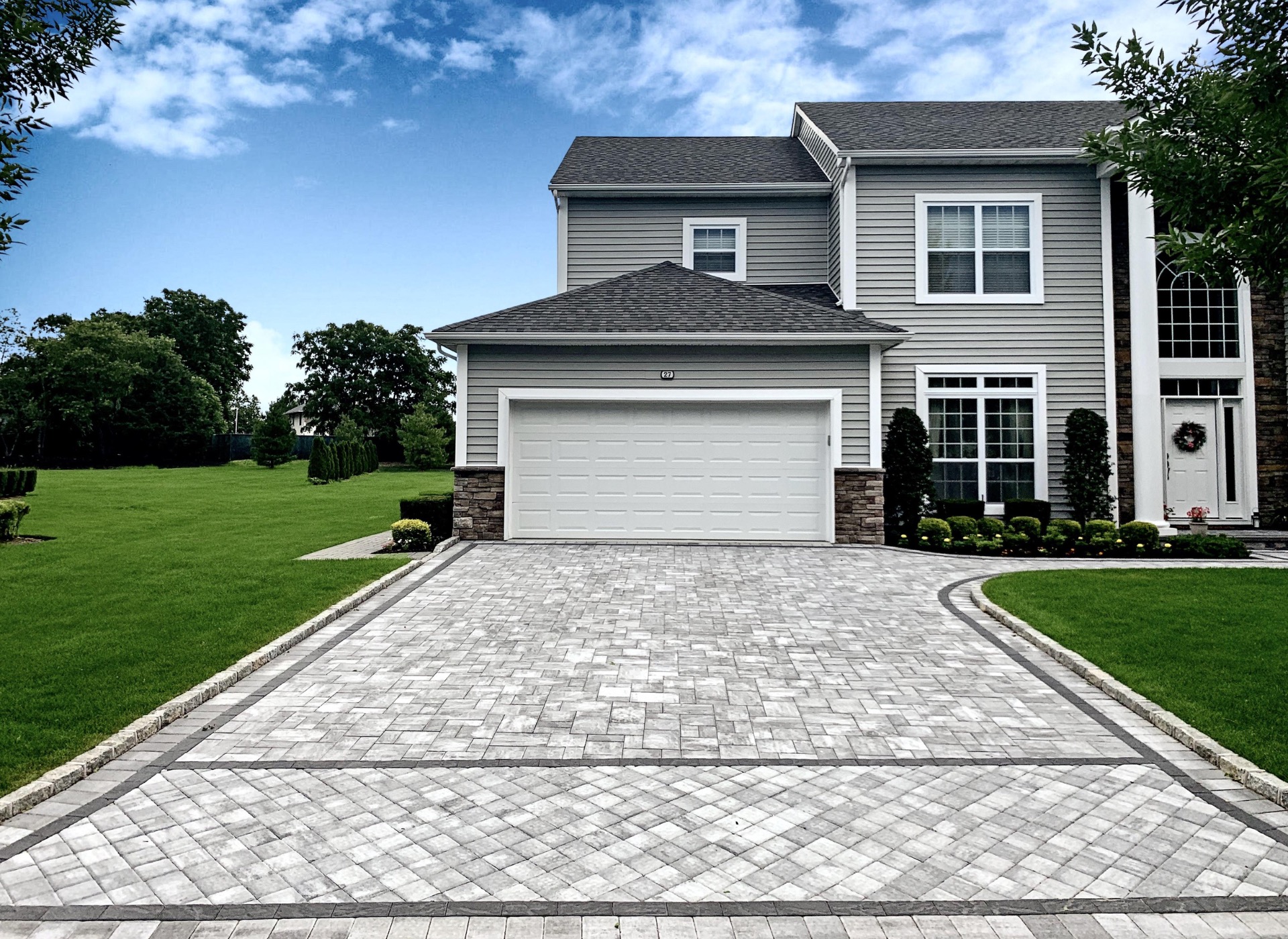 A two-story house with gray siding and a double garage sits at the end of a long, paved driveway. The lawn is well-manicured, and the sky is partly cloudy.