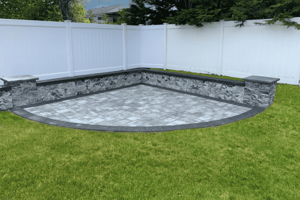 Stone patio with gray tiles and a curved edge, bordered by stone walls and white fencing, is situated on green grass under a cloudy sky.