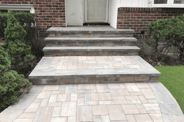 Stone steps and a pathway lead to a door of a brick building, bordered by trimmed bushes and grass.
