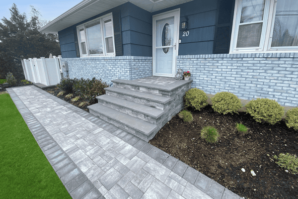 Brick house with gray stone walkway and steps leading to a pale blue door, surrounded by neatly trimmed shrubs and a lawn.