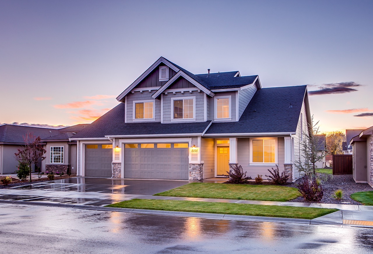 Suburban two-story house with a double garage and landscaped yard at dusk, wet driveway reflecting the pink sky.