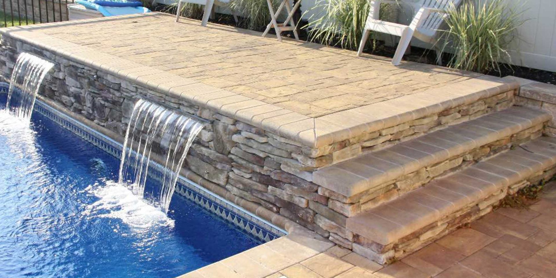 Pool area with stone steps and a waterfall feature flowing into the blue water. Two white chairs are placed on the paved surface above the steps.