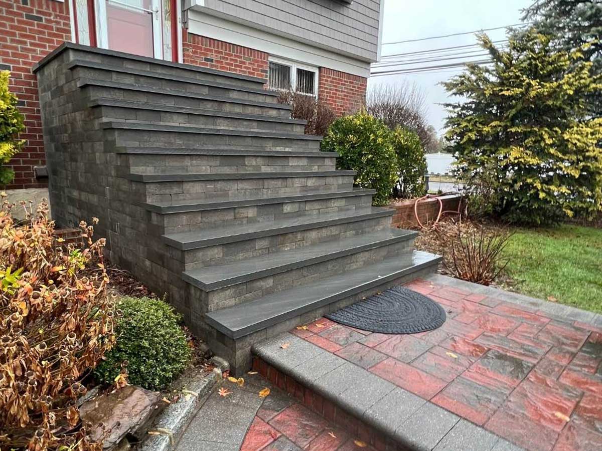 Gray stone steps lead up to the door of a brick house, surrounded by bushes and a wet pavement area.