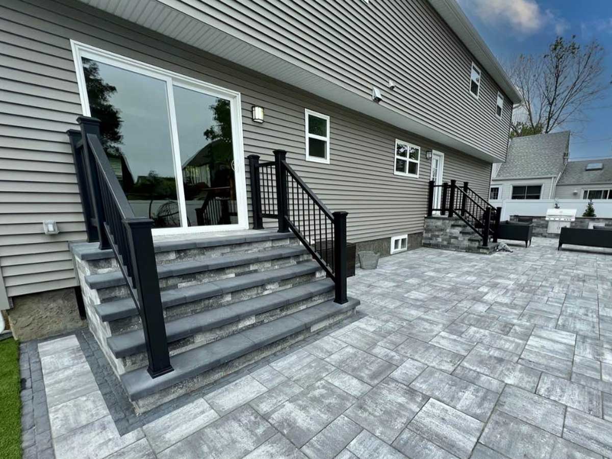 A house with gray siding has two sets of steps leading to a stone patio with a black railing. The patio features light gray tiles and has outdoor seating.