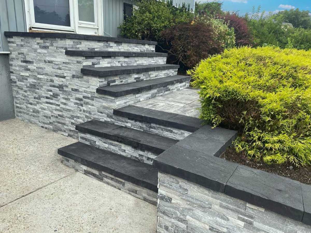 Stone steps and a small retaining wall with black edges lead up to a doorway, bordered by green shrubs and a concrete path.