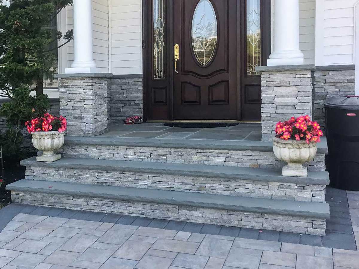 Stone front steps with two planters of pink flowers on either side, leading to a dark wooden door with decorative glass panels in a white house facade.