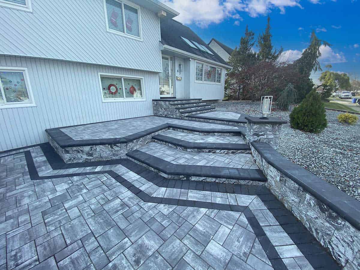 A modern house entrance with gray stone steps and paver walkway, featuring geometric patterns and a landscaped garden with shrubs.