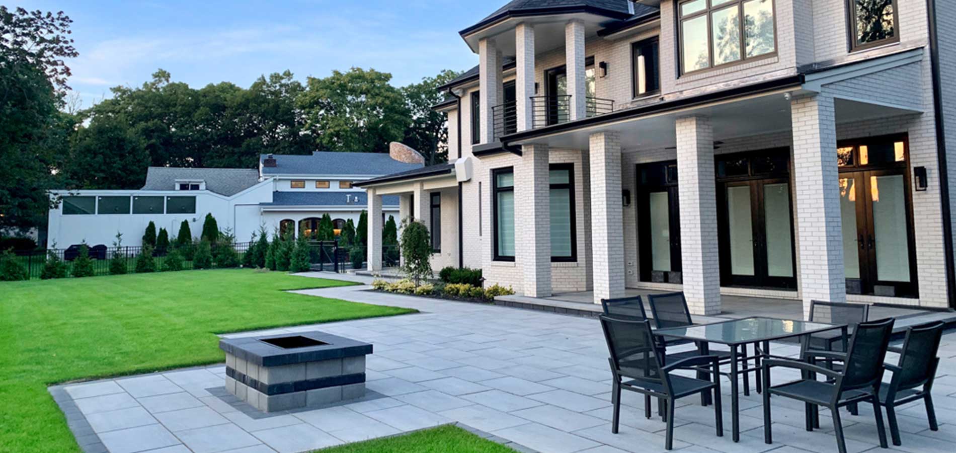 A large two-story house with white brick exterior and multiple archways, featuring a landscaped yard and a patio with dining area.