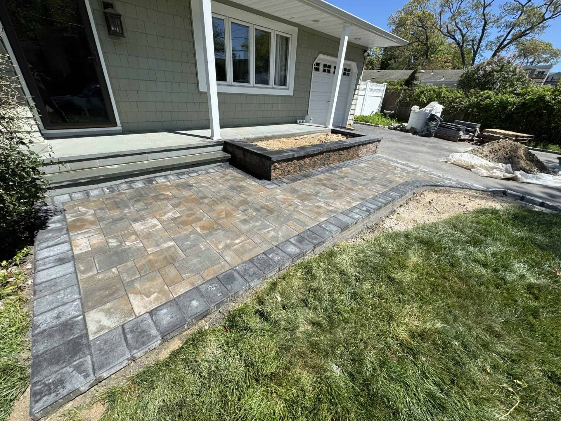 A newly constructed patio with gray and brown paving stones is adjacent to a house. Nearby is a rectangular planter and a grassy lawn partially under construction.