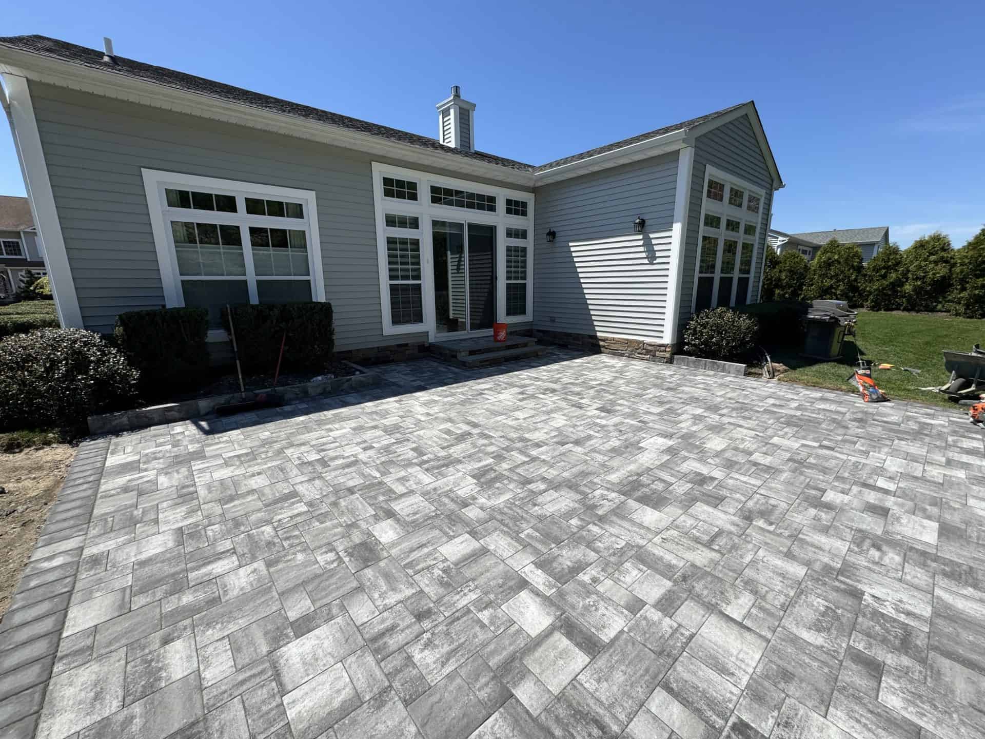 A house with gray siding features a newly laid stone patio with a herringbone pattern. Bushes line the patio edges, and gardening tools are visible. Clear blue sky in the background.