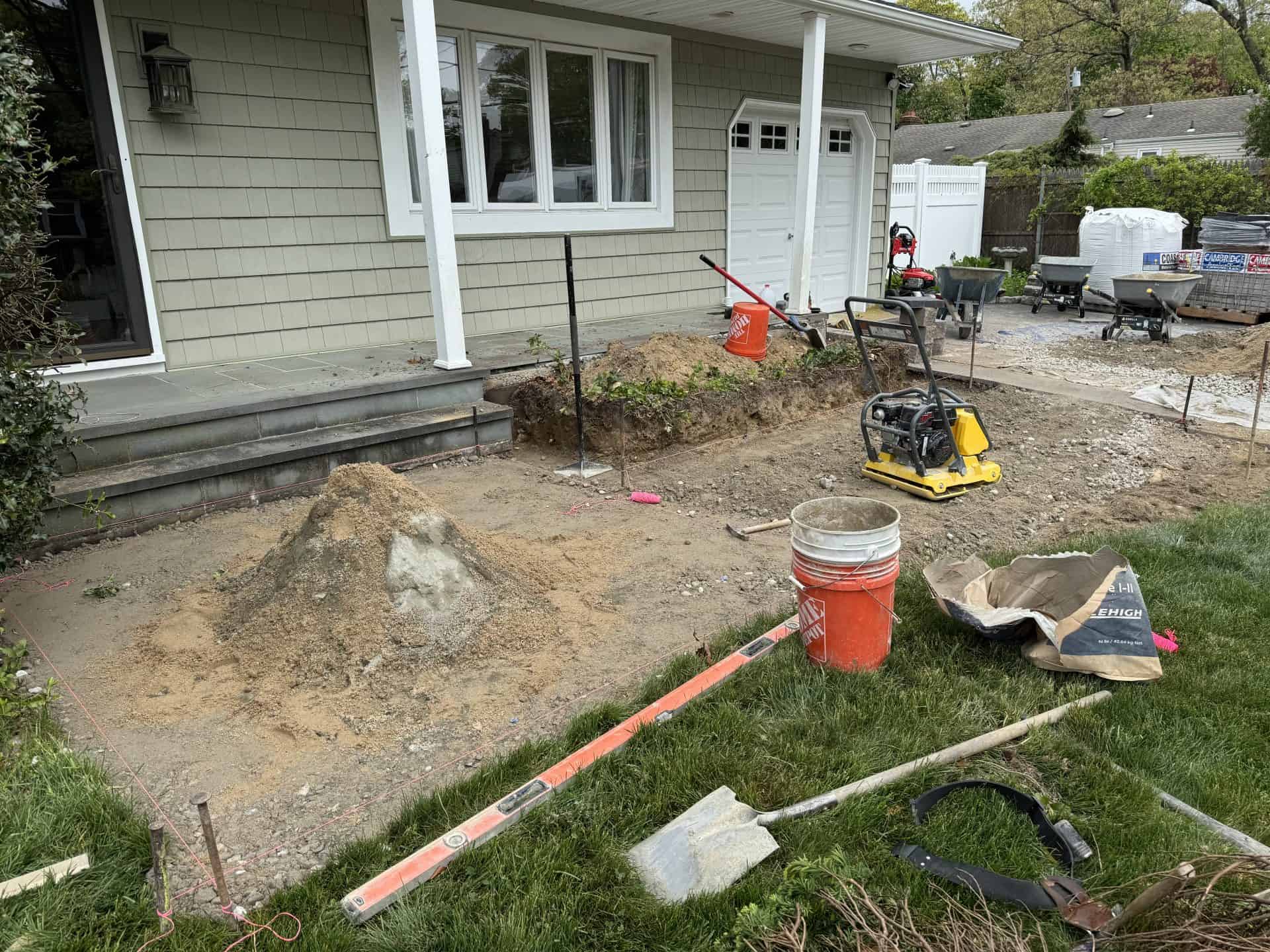 Residential front yard under construction with tools, equipment, and materials scattered. A mound of dirt is beside the steps to the entrance, and a compact plate compactor is visible.