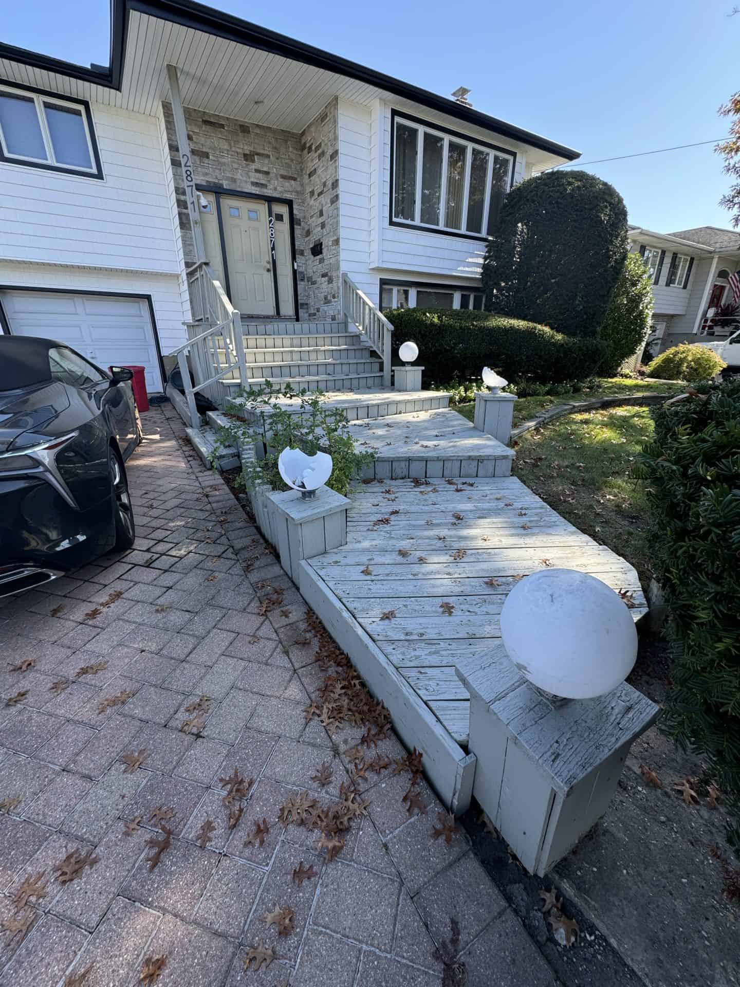 Ramp with wooden planks leading to a house entrance. White spherical lights line the path. The house has gray bricks and white siding, with shrubs nearby. A car is parked to the left.