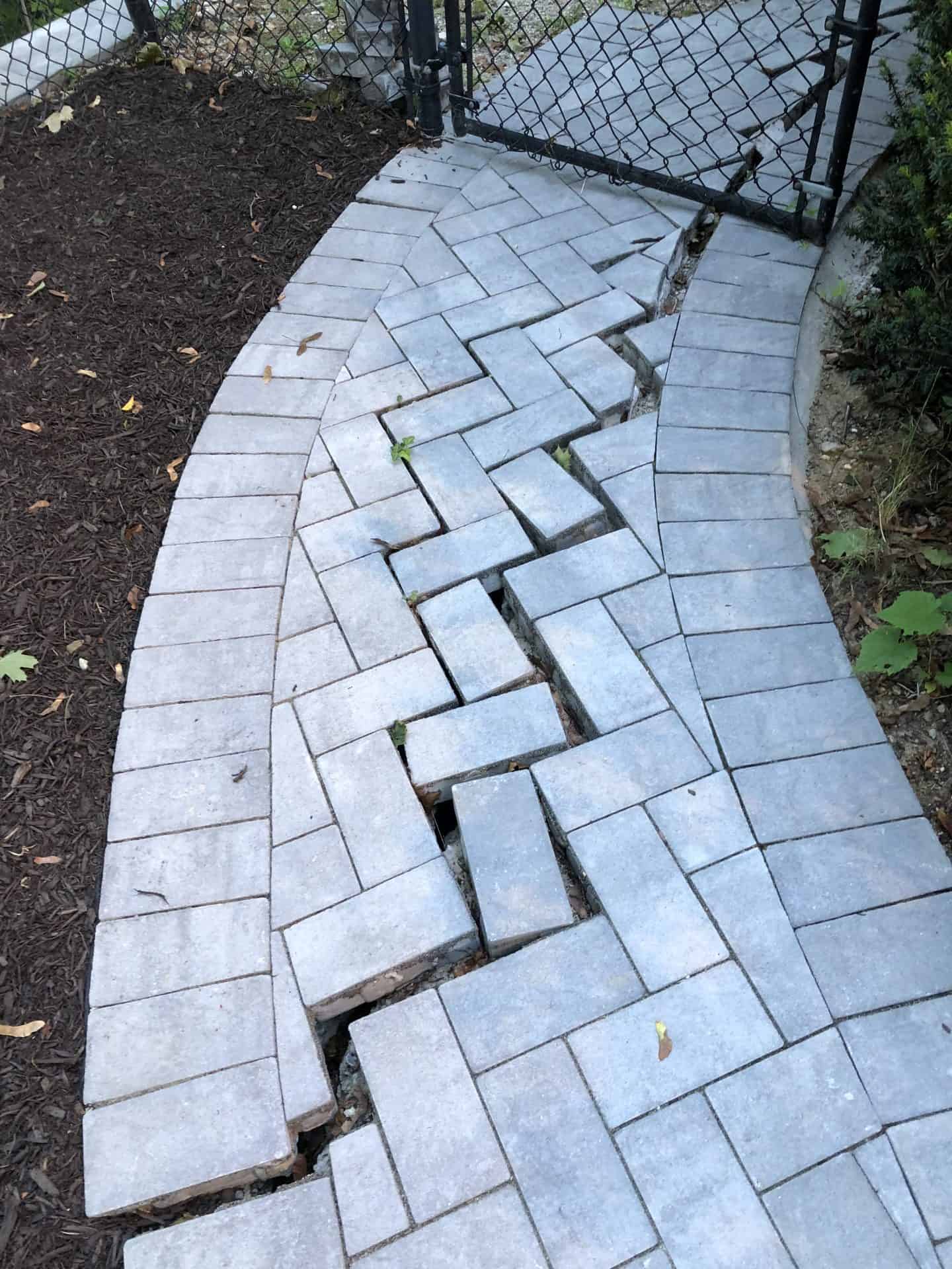 A paved walkway with gray bricks, some of which are uneven and shifted, leading to a black metal gate.