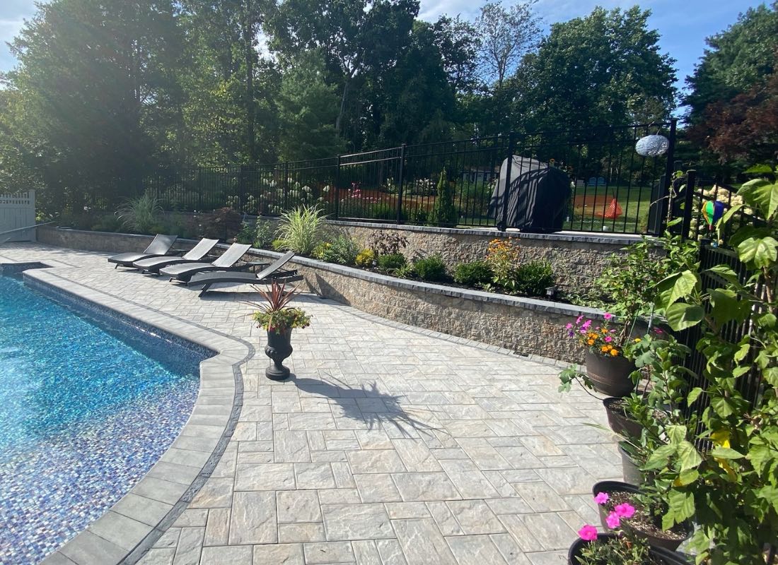 Poolside patio featuring four lounge chairs, potted plants, and a beautiful stone retaining wall that echoes the quality of Retaining Walls Baldwin. A grill and lush grassy area peek behind the fence, while trees and shrubs enhance this tranquil setting.