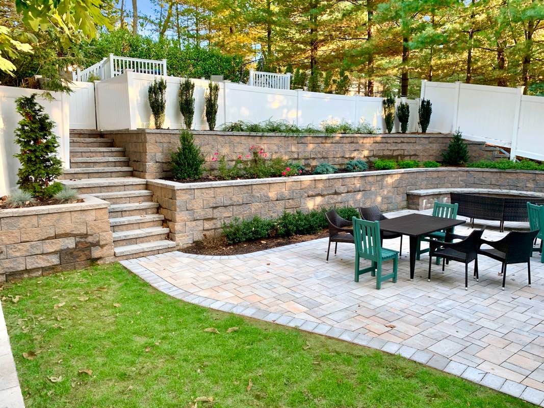 Landscaped backyard with stone walls and steps leading to an elevated garden area, flanked by trees and featuring outdoor dining furniture on a paved patio.