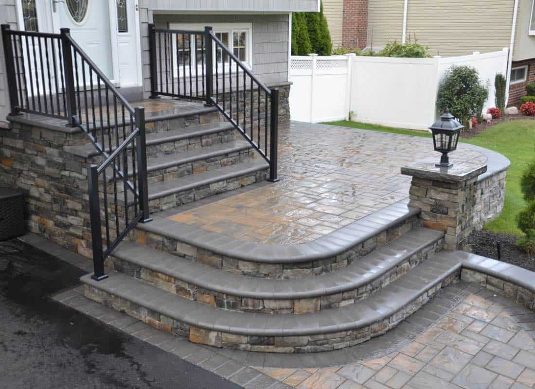 Stone steps with black railings lead to a front door. The patio features a stone wall with a decorative lamp post.