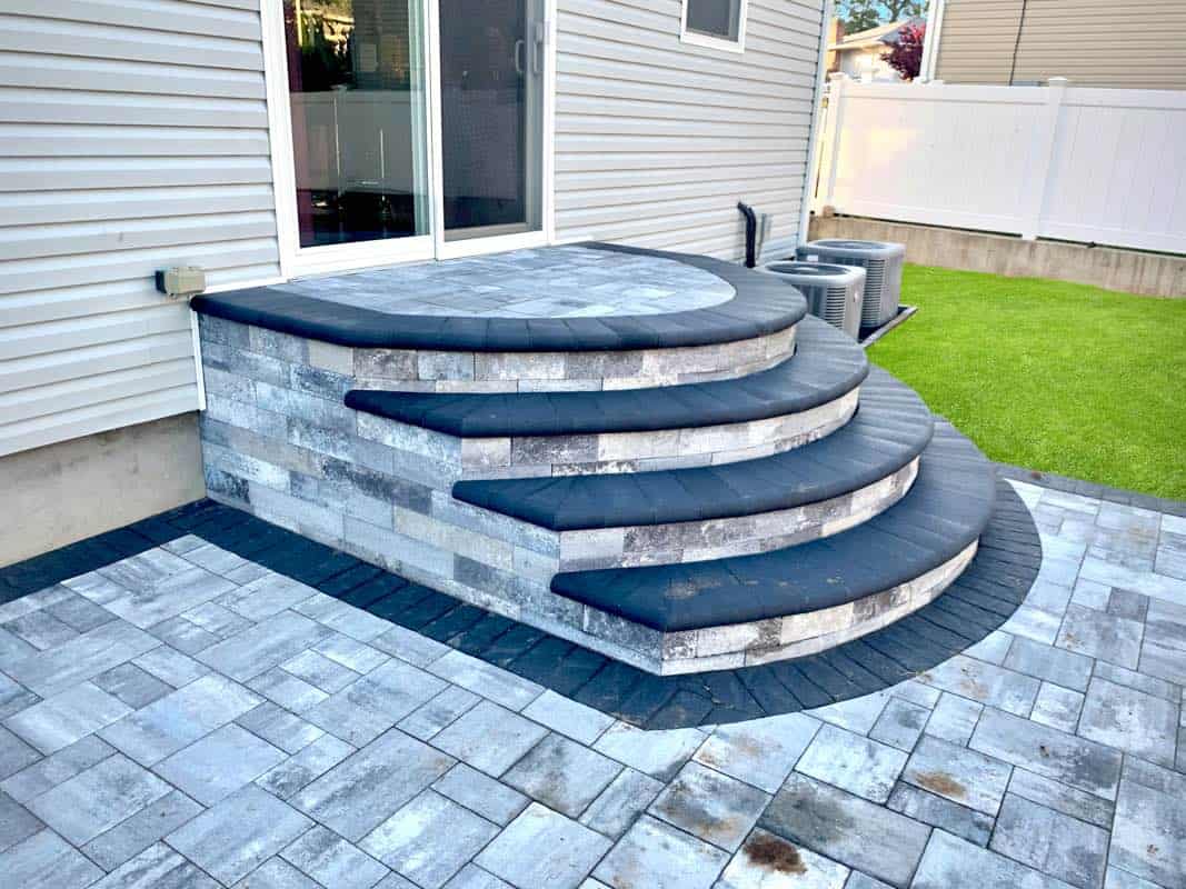 A curved stone patio steps up to a house with a sliding door. The steps have a dark stone cap, and the surrounding area is paved with a matching pattern. A green lawn and white fence are visible.