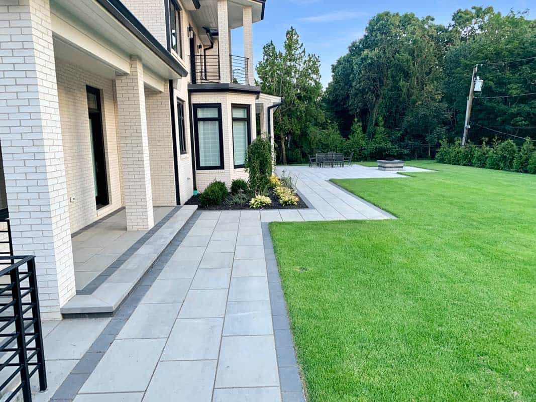 A modern house with white brick walls features a spacious patio and a neatly manicured lawn, bordered by trees in the background.