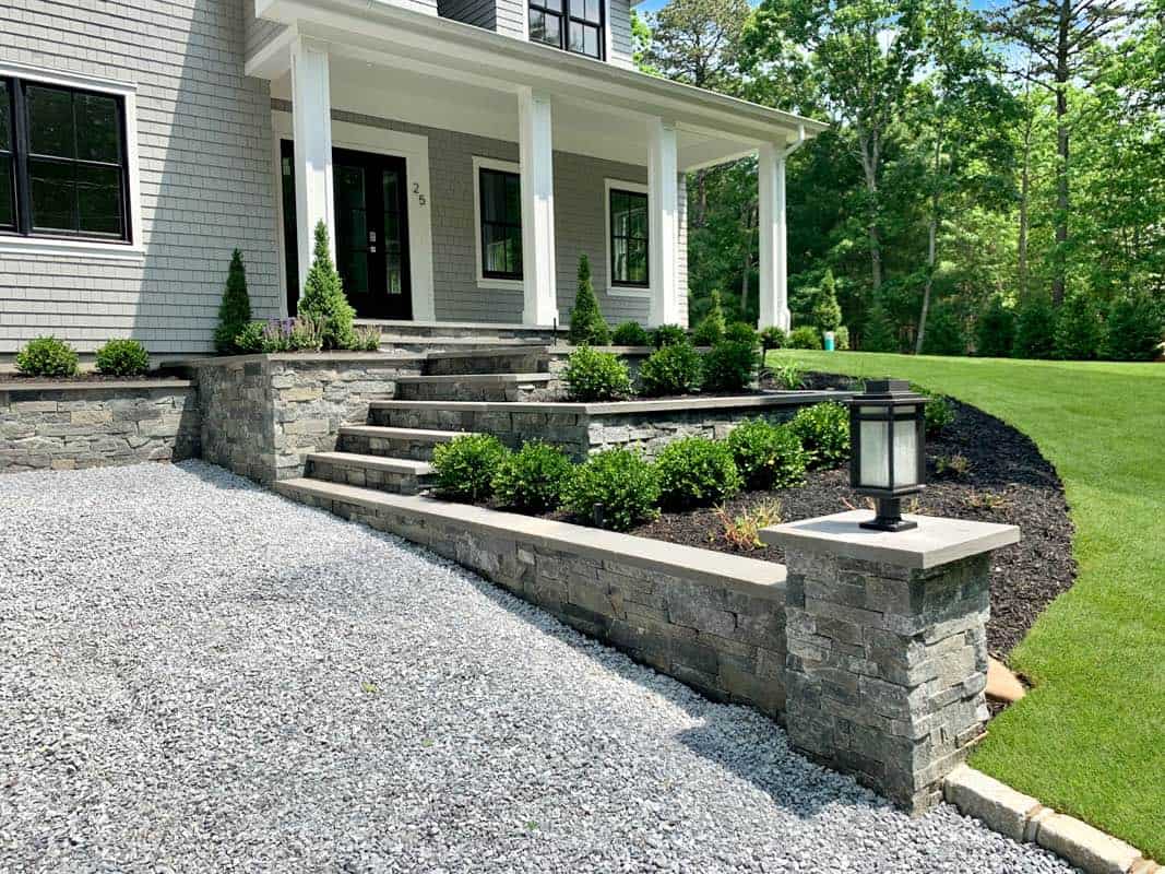 House exterior with a stone walkway, steps, and retaining wall. Neat shrubs line the path. The house features a covered porch with columns and shingle siding, set beside a well-maintained lawn.