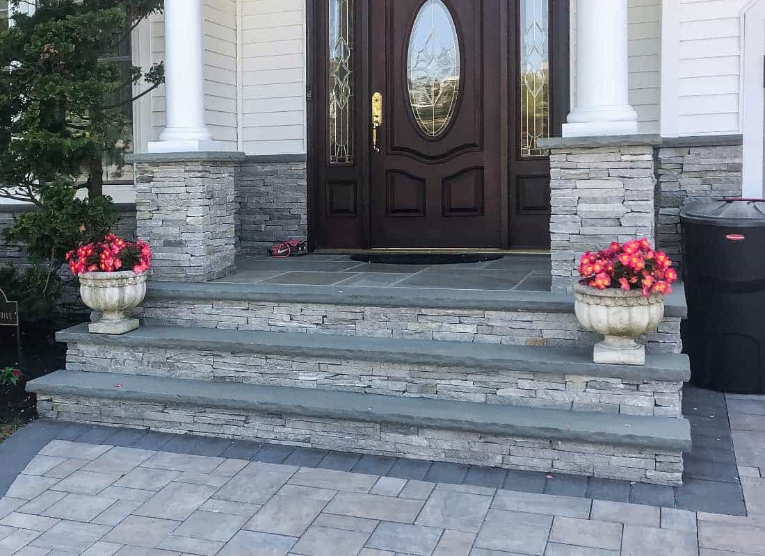 A front entrance with stone steps, two white columns, a dark wooden door with glass, and stone planters with pink flowers on each side. A small hedge is partially visible on the left.
