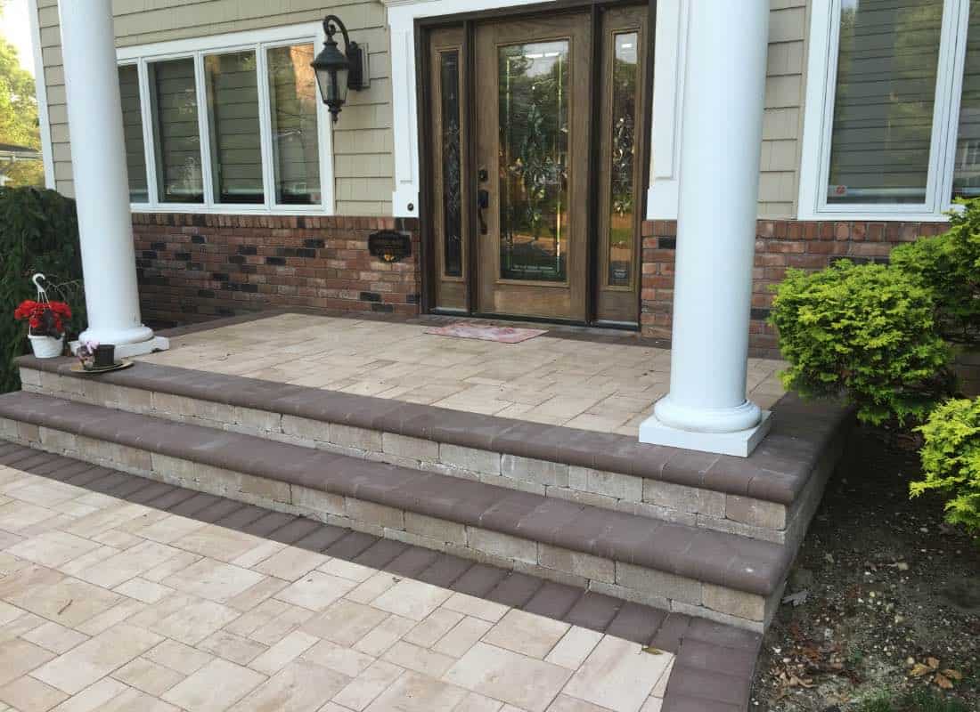Front porch with a tiled staircase, two white columns, a brick wall, and a wooden door with glass panels. A small plant and red flowers are on the steps.