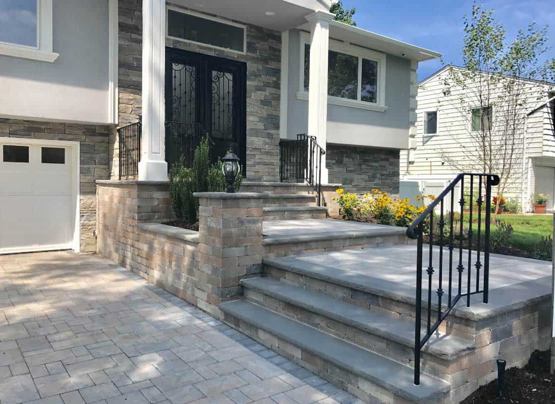 Front entrance of a house with stone steps, black railing, and a flower bed to the right. White columns support a small porch roof.