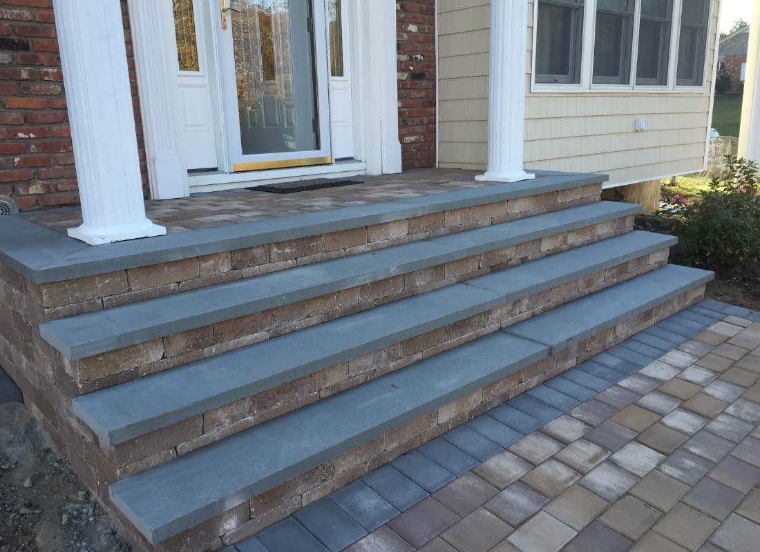 Brick and stone steps leading to a porch, with a paved pathway in front and a side view of a house.
