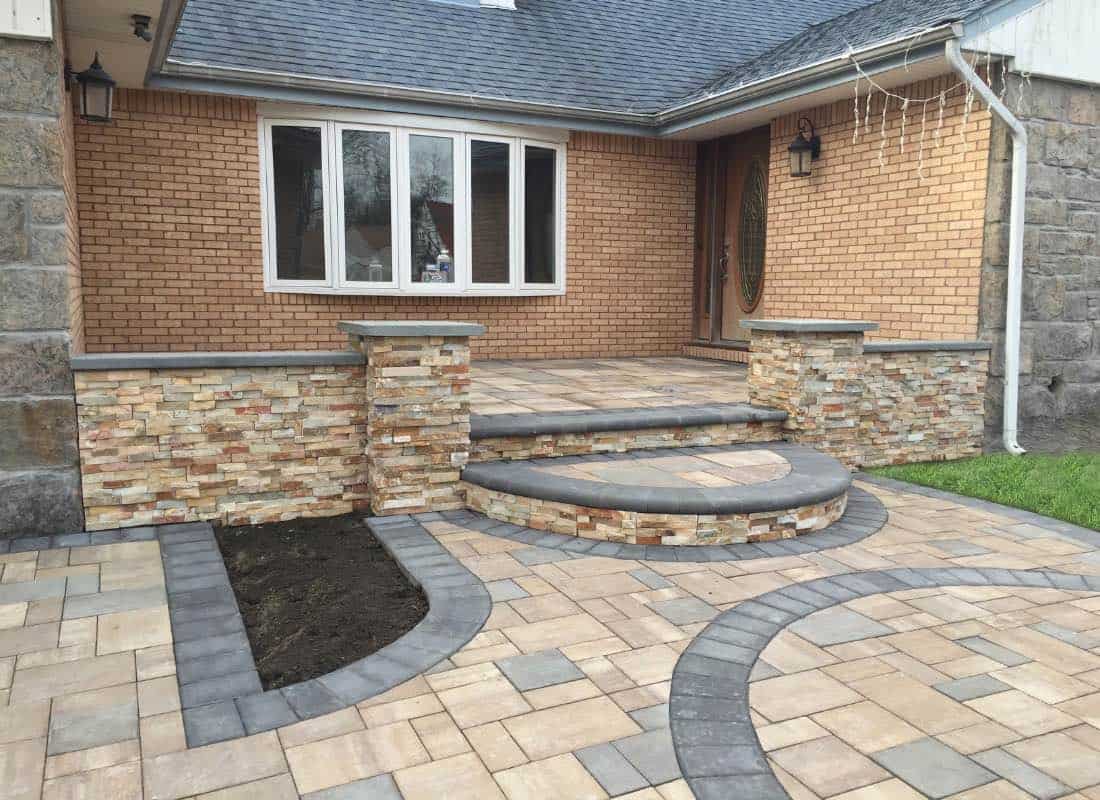 A house entrance with a brick facade features a paved walkway, stone steps, and small pillars flanking the door.