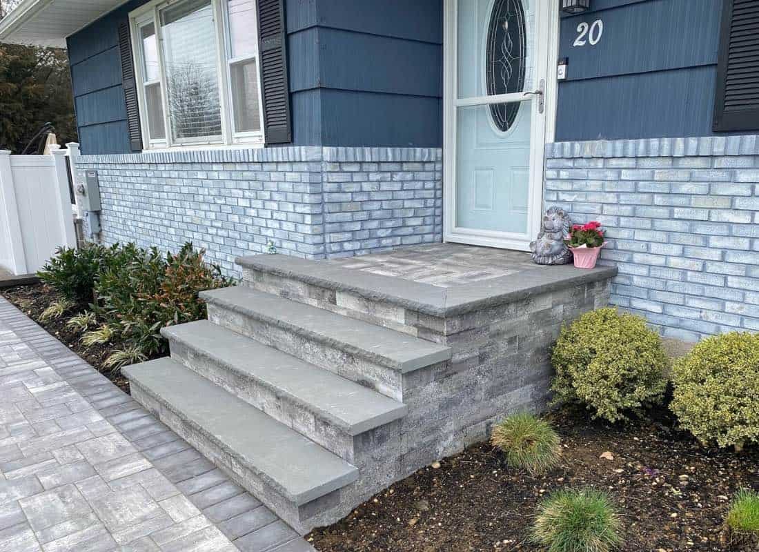 Brick house entrance with gray stone steps and a walkway, featuring a potted pink flower and a decorative figure on the step. Bushes and small plants line the path.