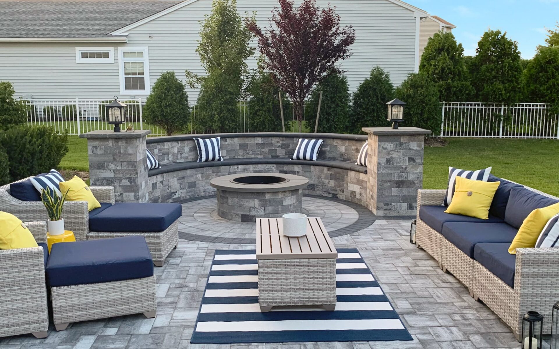 Outdoor patio with wicker furniture, a circular stone fire pit, striped cushions, and a striped rug. Surrounded by greenery and set against a residential backdrop.