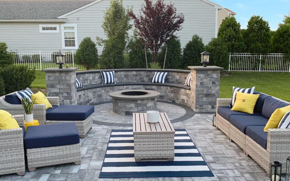 Outdoor patio with wicker furniture, a circular stone fire pit, striped cushions, and a striped rug. Surrounded by greenery and set against a residential backdrop.