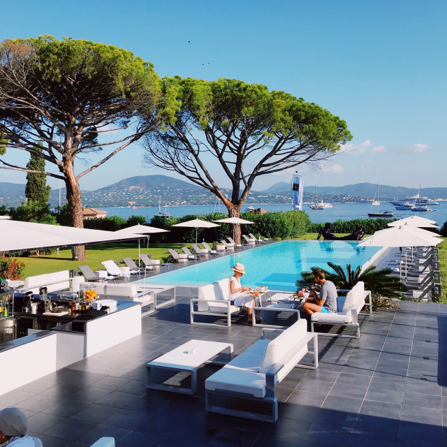Two people dining by an outdoor pool overlooking a bay with yachts and mountains in the background, sunny day with clear skies and large trees.
